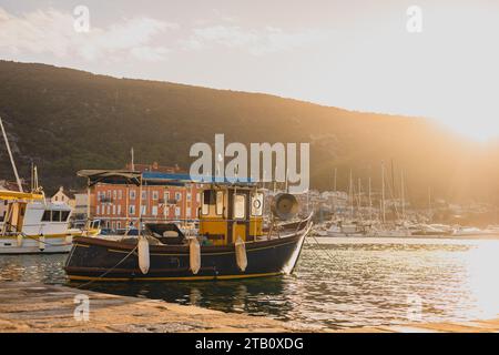 Barca da pesca ormeggiata nelle prime ore del mattino nel porto di Cherso. Una bella barca da pesca sul mare calmo in un porto. Il sole sorge lentamente dietro una collina. Foto Stock