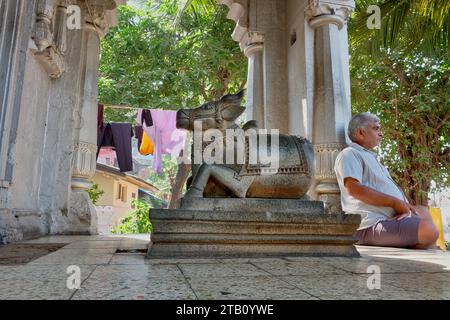 Un uomo siede accanto alla figura di un toro Nandi, il veicolo del dio Shiva, in un piccolo tempio Shiva a Banganga Tank, Walkeshwar, Mumbai, India Foto Stock