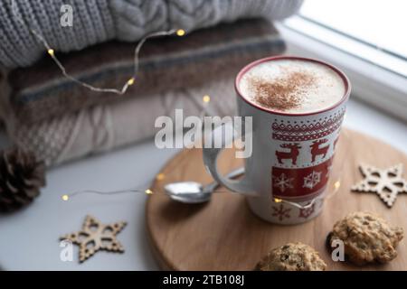 Natura morta del davanzale invernale. Tazza di caffè caldo in ceramica rossa sul davanzale. Decorazioni natalizie sullo sfondo. Foto di casa. Lana calda kn Foto Stock