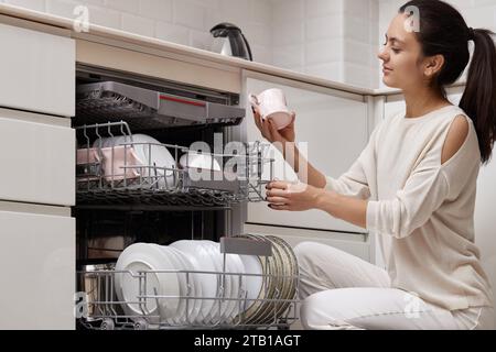 una donna attraente che scarica la tazza dalla lavastoviglie automatica aperta con utensili puliti all'interno in una moderna cucina bianca. Foto Stock