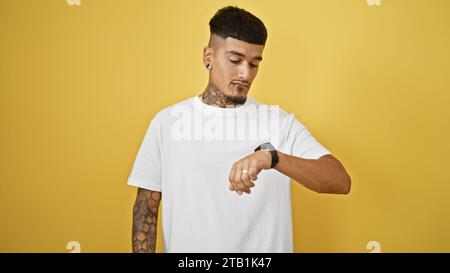 Bellissimo giovane latino, tatuato e rilassato, guardando il suo orologio da polso con una seria espressione. e' in piedi contro un bac giallo isolato e figo Foto Stock