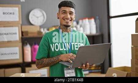 Bellissimo giovane latino, tatuato e indossato in t-shirt, sorridendo con sicurezza mentre si offre volontario con un portatile al centro di beneficenza, al servizio della comunità con un Foto Stock
