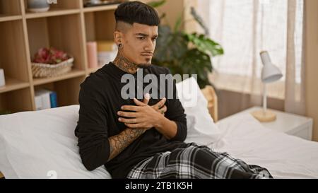 Bel giovane latino, tatuato e sofferente, seduto sul suo letto che stringe il petto in agonia per attacco di cuore a casa Foto Stock