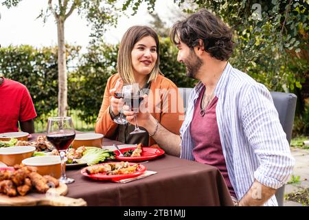 Una coppia si impegna in una conversazione amichevole con il vino durante un evento all'aperto, con un tavolo pieno di vari piatti in un lussureggiante giardino. Foto Stock