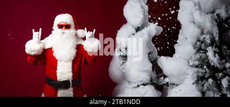 Foto banner ravvicinata di Babbo Natale alla moda con occhiali da sole posa e si diverte su sfondo rosso con alberi di natale Foto Stock