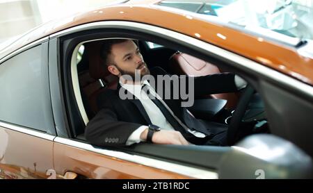 Ritratto ravvicinato di un uomo d'affari bello, sicuro di sé, elegante e barbuto mentre guida la sua auto. Auto elettrica di lusso moderna Foto Stock