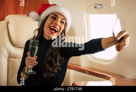 Ritratto ravvicinato di una bella bruna sorridente nel cappello di Babbo Natale con un bicchiere di champagne in mano, scatta selfie sul suo smartphone mentre vola in volo Foto Stock