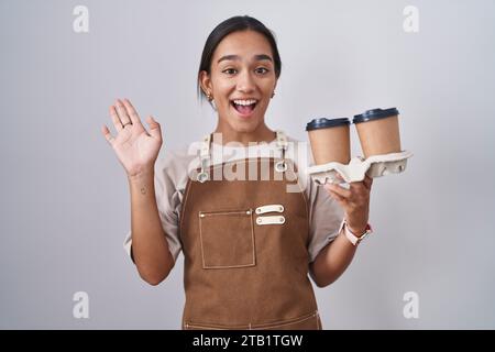 Giovane donna ispanica che indossa grembiule da cameriera professionale che tiene in mano il caffè, salutando con un gesto di benvenuto felice e sorridente Foto Stock