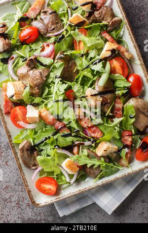 Insalata calda con fegato di pollo grigliato, pancetta, pomodori ciliegini, rucola, cipolla rossa e crostini da vicino sul piatto sul tavolo. Vista dall'alto verticale da abo Foto Stock