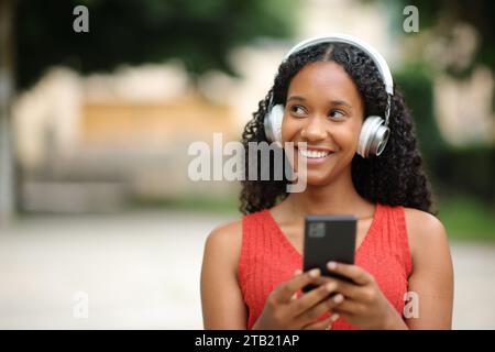 Il ritratto frontale di una donna nera felice guarda di lato ascoltando la musica che cammina per strada Foto Stock