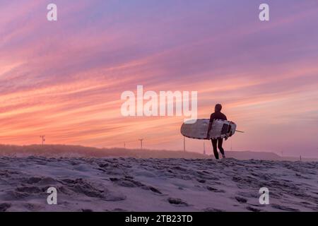 Un surfista cammina sulla spiaggia con la sua tavola da surf al tramonto Foto Stock