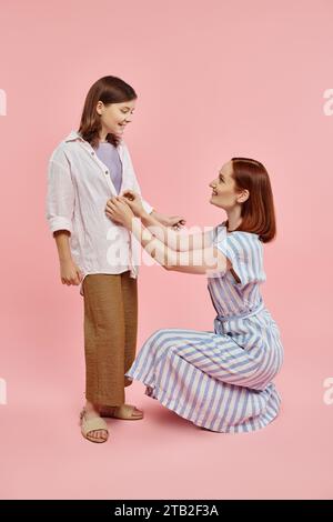 madre seduta su fianchi e camicia da abbottonatura di una preadolescente su sfondo rosa, amore e cura Foto Stock