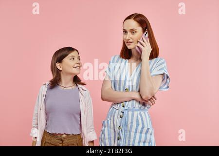 donna alla moda che parla al telefono cellulare vicino a una preadolescente sorridente su sfondo rosa, mamma e adolescente moderne Foto Stock