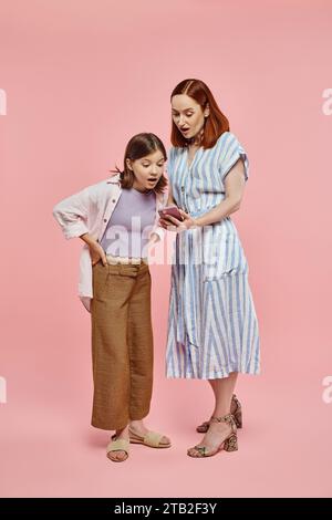 la madre e la figlia adolescente hanno stupito guardare il telefono cellulare mentre sono in piedi su uno sfondo rosa Foto Stock