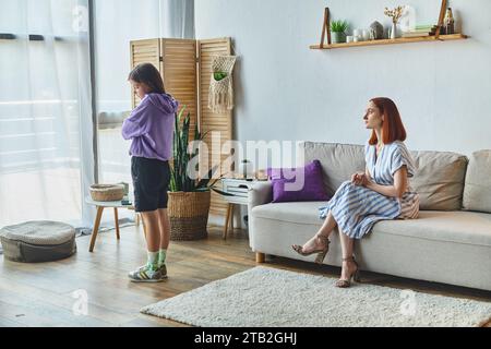 ragazza adolescente offesa in piedi con le braccia piegate vicino a madre scontenta sul divano, conflitto familiare Foto Stock