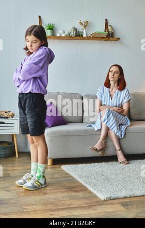 ragazza adolescente offesa in piedi con le braccia piegate vicino a madre scontenta sul divano, conflitto familiare Foto Stock