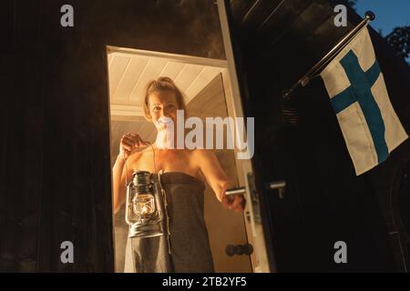 Donna sorridente in un asciugamano con una lanterna all'ingresso di una sauna finlandese mobile a botte al crepuscolo. Foto Stock