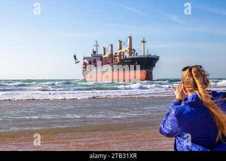 Una donna filma sul suo telefono come un kitesurfer su una tavola vola in aria vicino a una grande nave in mare. Vityazevo, Russia - 12.02.2023 Foto Stock