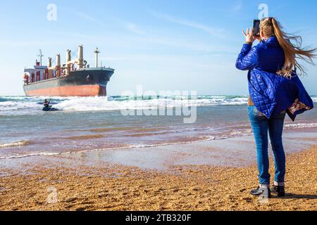 il turista filma sul suo telefono come un kite surfista naviga sulle onde vicino a una grande nave da carico ancorata sulla costa. Foto Stock