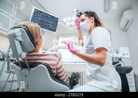 Paziente clinica odontoiatrica visita moderno reparto medico. Medico dentista vestita uniforme con lenti di ingrandimento che puntano la luce. Paziente seduto sulla sedia. Foto Stock