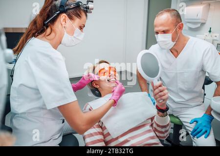Donna sorridente sdraiata in sedia di stomatologia, guardando allo specchio, medico femminile con escavatore e specchio strumenti medici e assistente uomo dopo denti whit Foto Stock