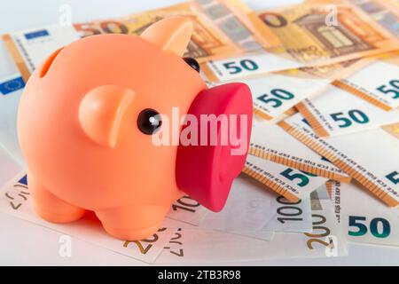 Un salvadanaio rosa si trova su un tavolo coperto da una grande quantità di banconote in euro su uno sfondo chiaro da vicino Foto Stock