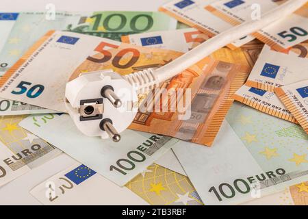 Una spina elettrica bianca si trova su un tavolo coperto da una grande quantità di banconote in euro su uno sfondo chiaro da vicino Foto Stock