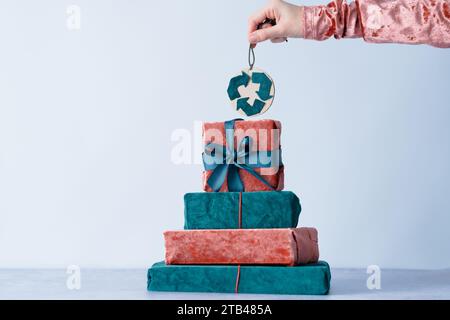Regali di Natale. Composizione con confezione regalo in tessuto ecologico sotto forma di albero di Natale e mano di un bambino che tiene un simbolo di riciclaggio. Foto Stock