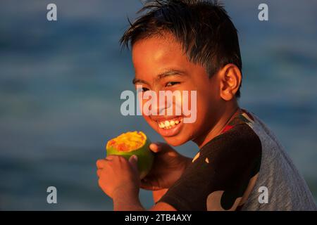 Ragazzo locale che mangia un mango maturo sul molo della città in questa isola di vacanza circondata da coralli e meta per immersioni subacquee. Isola di Bunaken, Sulawesi, Indonesia Foto Stock