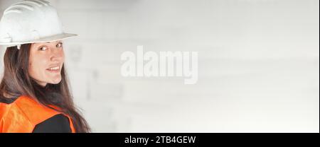 Giovane donna in elmetto bianco e giubbotto arancione ad alta visibilità, capelli lunghi e scuri, guardando sopra la spalla nella macchina fotografica, sorridendo. Costruzione sfocata si Foto Stock