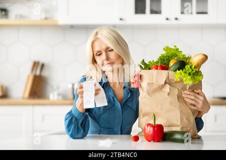 Una casalinga anziana confusa che controlla le bollette di alimentari in cucina dopo aver fatto acquisti di cibo Foto Stock
