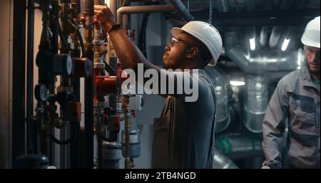 I lavoratori dell'industria pesante afroamericana che indossano uniformi di sicurezza e elmetto ispezionano il sistema di tubazioni nei moderni impianti o impianti energetici industriali. Un ingegnere professionista gestisce la fabbrica di produzione. Foto Stock