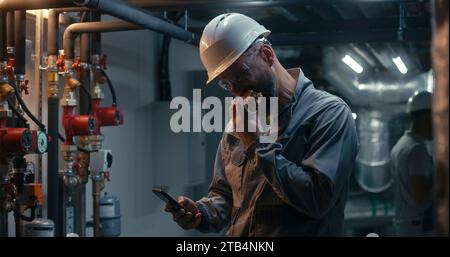 Un ingegnere professionista lavora in una fabbrica moderna, mangia hamburger e usa il telefono. In background un collega afroamericano ispeziona il sistema di tubazioni utilizzando un tablet. Concetto di impianto di produzione industriale. Foto Stock