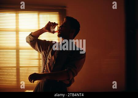 Giovane in abbigliamento retrò che beve whisky davanti alla macchina fotografica mentre lavora fino a tardi in un ufficio buio con finestra coperta da veneziane Foto Stock