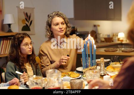 Donna ebrea matura che accende le candele sul candelabro della menorah al tavolo servito mentre si siede accanto a sua figlia e si prepara per la cena Foto Stock