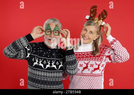 Felice coppia anziana con maglioni natalizi, cerchietto da renna e divertenti occhiali su sfondo rosso Foto Stock