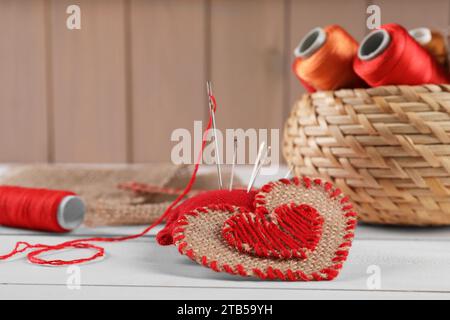 Cuore di tessuto burlap con punti rossi e bobine di fili su un tavolo di legno bianco. Spazio per il testo Foto Stock