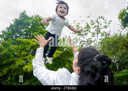 Bella giovane mamma indiana che lancia la bambina in aria e cattura, diverte e si diverte insieme nel parco. Foto Stock
