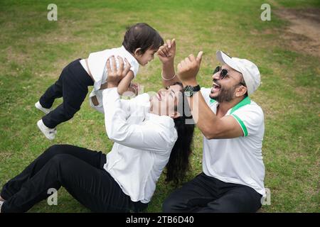 Giovane famiglia indiana di tre mamme che lancia la bambina in aria e papà diverte la bambina e si diverte insieme nel parco. Foto Stock