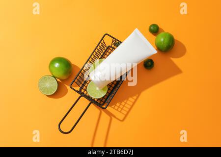 Primo piano di una provetta cosmetica senza etichetta posta in un cestello a rete metallica con limone fresco su sfondo arancione. Il limone contiene acido L-ascorbico, che ha Foto Stock