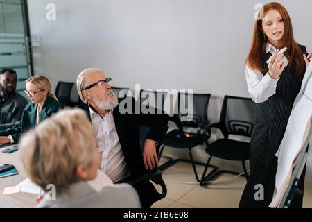 La vista laterale di una giovane donna d'affari presenta un nuovo progetto su una lavagna a fogli mobili per dipendenti di diverse età, sia femminili che maschili. Foto Stock