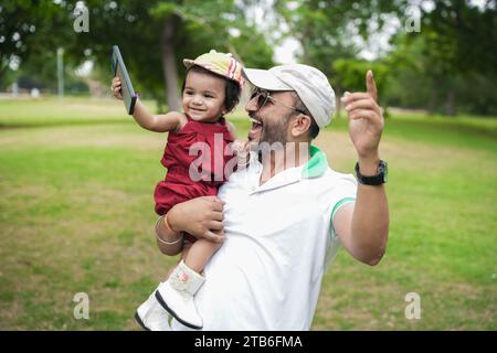 Bellissimo giovane padre indiano che indossa cappello e occhiali da sole e danza con la sua bambina nel parco o in giardino. Foto Stock