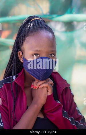 villaggio, giovane ragazza africana con lunghe trecce, che indossa una maschera tessile, sfondo verde all'aperto Foto Stock