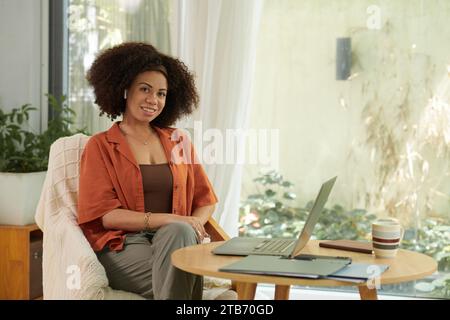 Ritratto di una donna sorridente seduta su un tavolino da caffè con un computer portatile aperto, che lavora da casa Foto Stock