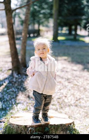 Una bambina sorridente con le mani nelle tasche del gilet si trova su un ceppo nel parco Foto Stock