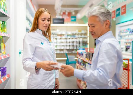 Giovane farmacista donna in uniforme bianca che assiste il cliente anziano di sesso maschile nell'acquisto e nella scelta dei farmaci dagli scaffali del negozio Foto Stock