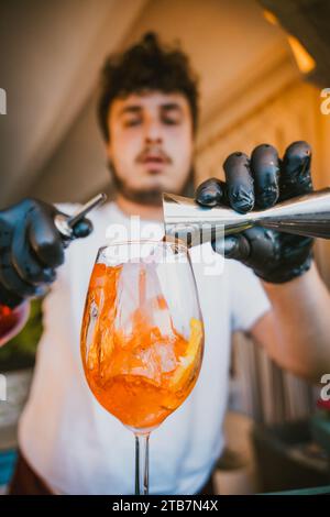 Giovane barman professionista concentrato che versa una bevanda all'arancia da jigger in un bicchiere mentre prepara un cocktail rinfrescante e si trova al bancone del ba Foto Stock
