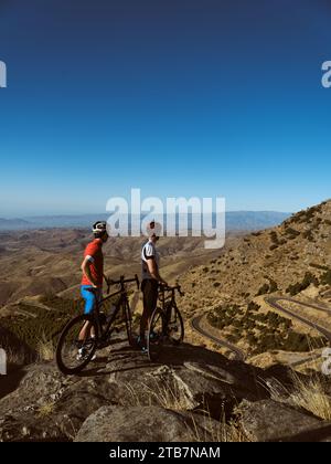 Vista laterale due ciclisti in piedi con le loro mountain bike che si affacciano su una strada tortuosa e colline ondulate Foto Stock