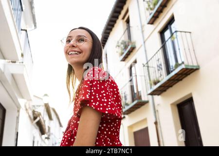 Vista dall'angolo basso di una giovane turista allegra che indossa casuali e occhiali da vista sorridente e che guarda lontano mentre si trova in piedi contro vecchi edifici Foto Stock