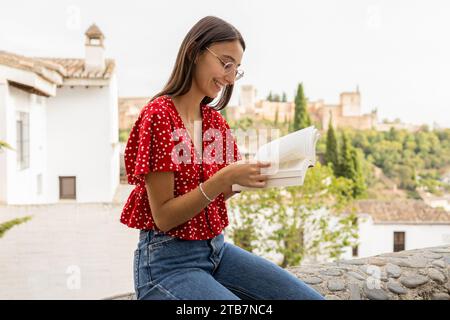 Giovane donna felice e concentrata che indossa casuali e occhiali da vista leggendo un libro mentre è seduta su un muro di contenimento con la fortezza di Alcazaba e il cielo limpido nel backgrou Foto Stock